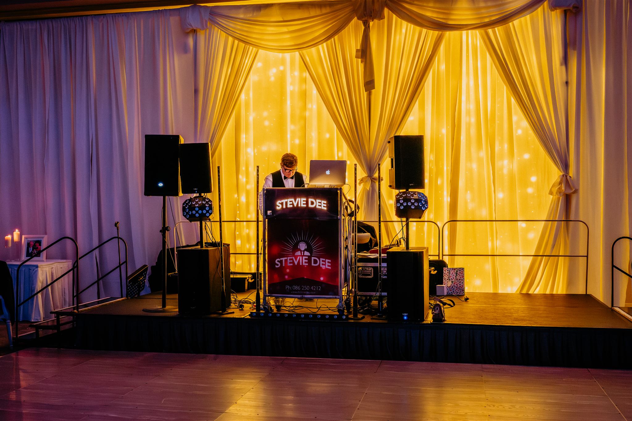 DJ booth and lighting rig in front of a gold draped wedding backdrop